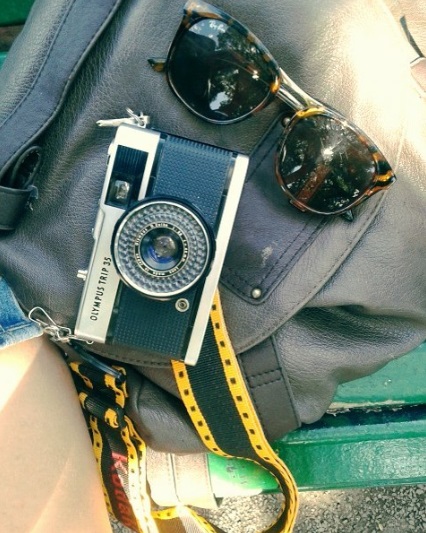 Cámara de fotos antigua junto a unos lentes de sol, sobre una cartera  de cuero negra.