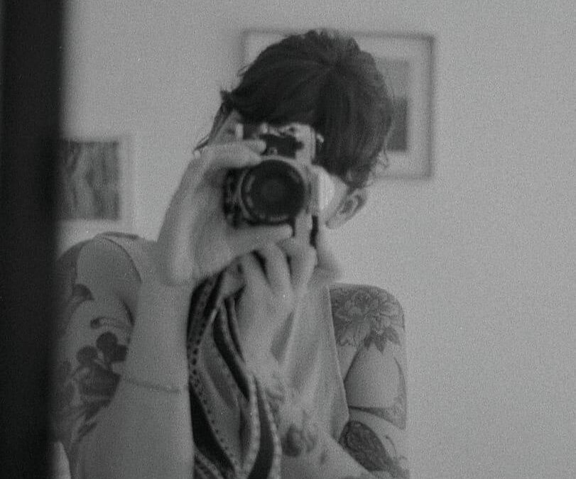 Mujer tomando un fotografia, ella tiene los brazos tatuado, imagen en blanco y negro.