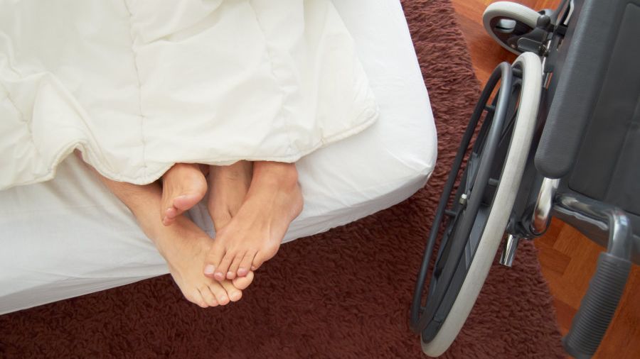 cuatro pies asoman sobre una cama con sábanas blancas. Al costado una silla de ruedas.