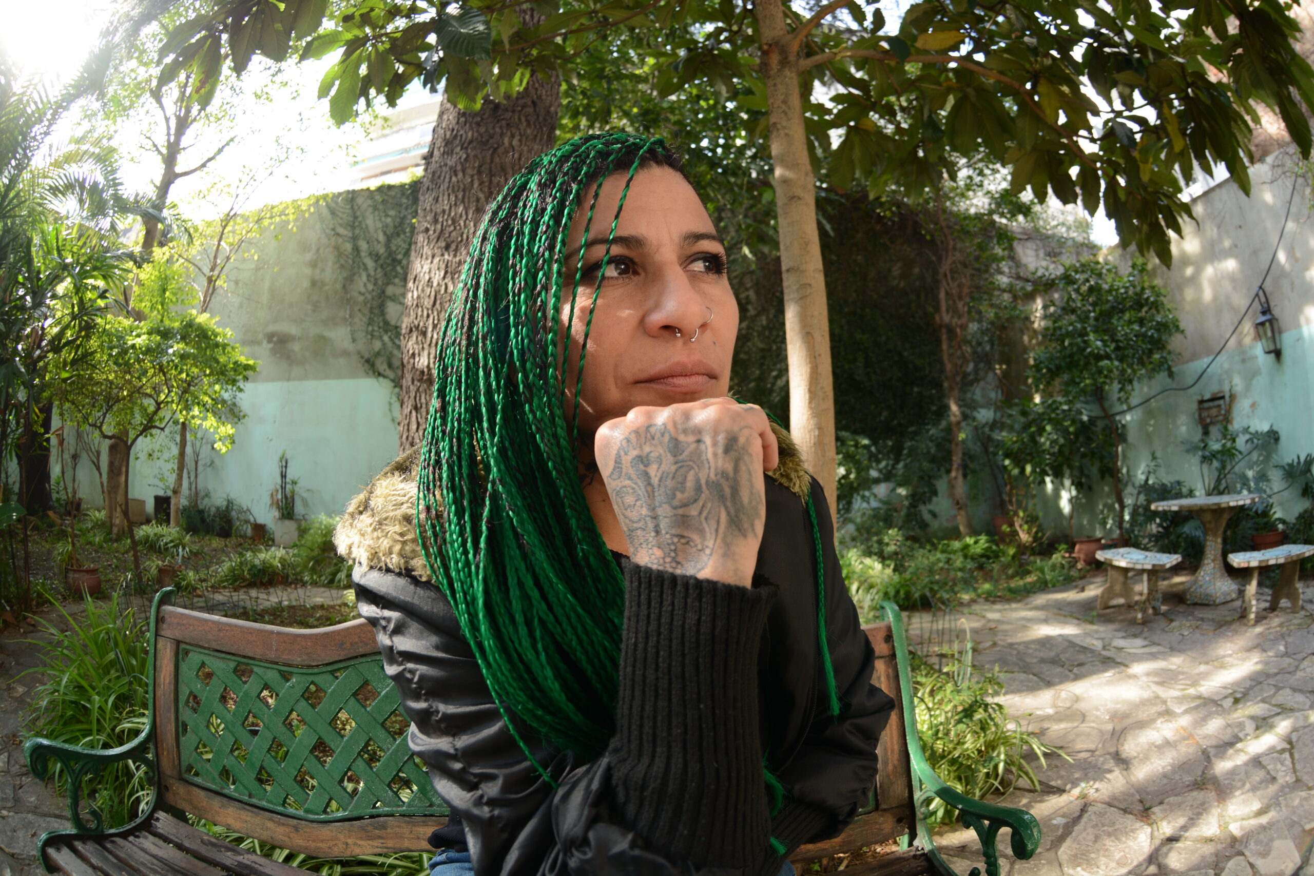 Caryna Moreno, sentada en el banco de un jardín, con una de sus manos apoyadas sobre la pera y una mirada pensativa. Tiene rastas de color verde en el pelo