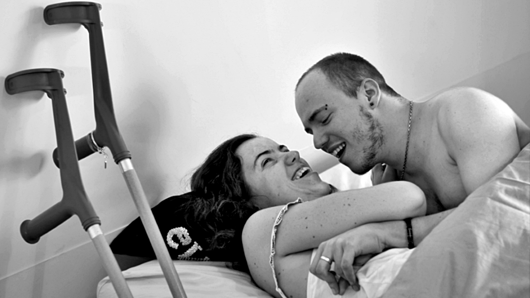 mujer y hombre abrazados en una cama riéndose, sus miradas están conectadas. A un costado, sobre una pared, están apoyados un par de bastones canadienses. 